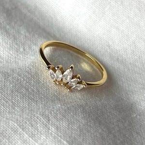 Può includere: Un anello color oro con un gruppo di pietre chiare a taglio marquise. La fascia dell'anello è sottile e liscia, e le pietre sono disposte in un motivo delicato e simmetrico. L'anello è posto su uno sfondo bianco.