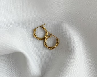 Puede incluir: Un par de pendientes de aro de oro con un diseño trenzado. Los pendientes están sobre un fondo de tela blanca.