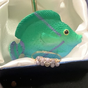 Puede incluir: Un anillo de oro con un diseño de alambre envuelto y cuentas de color lavanda. El anillo está sobre una superficie blanca delante de una figurita de pez verde y morado.
