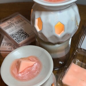 May include: Two white ceramic wax warmers with a pink wax melt in each. The wax warmers have a geometric design with open holes. The wax melts are in the shape of a triangle.
