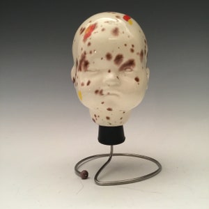 May include: A ceramic doll head with brown, yellow, and red paint splatter on a white background. The head is on a black stand with a metal wire base.