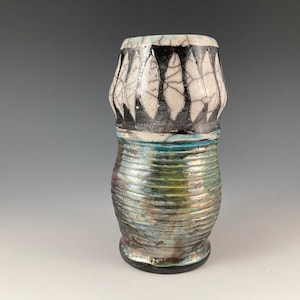May include: A handmade ceramic vase with a textured, ribbed body and a decorative top. The top features a black and white geometric design. The vase has a metallic, iridescent finish with shades of blue, green, and silver.