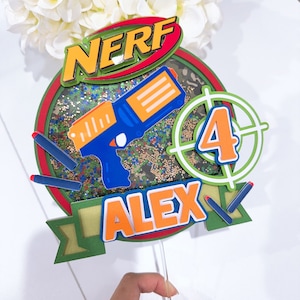 May include: A green, red and blue cake topper with the word "NERF" in a red and white circle, a blue and orange toy gun, a green target with the number "4" in the centre, and the name "ALEX" in orange letters.