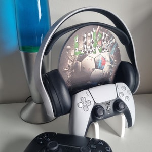 May include: A gaming setup featuring a white and black PlayStation 5 controller on a white stand, a black and white headset, and a decorative disc with a soccer ball design. A second black controller is visible.
