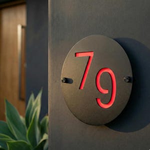 May include: A round, black metal address plaque with the number 79 cut out, revealing a red interior. The plaque is mounted on a dark gray wall with two small screws. A modern house number sign.