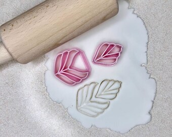 Könnte beinhalten: Drei Blätter-förmige Ausstecher auf einer weißen Oberfläche mit einem Holz-Nudelholz. Die Ausstecher sind rosa und weiß.