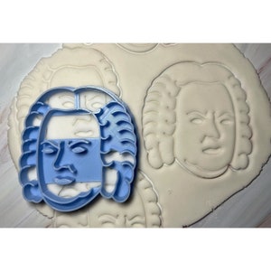 Op de afbeelding: Een blauwe plastic koekjesvormer in de vorm van een gezicht met een gepoederd pruik. De vormer ligt op een wit oppervlak met verschillende andere indrukken van de vormer.