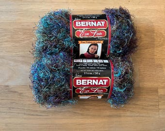 May include: Two skeins of Bernat Eye Lash Nouveau yarn in a variegated blend of dark purple, teal, and black fibers. Each label indicates a weight of 1 3/4 oz. / 50 g and a length of 77 yards. The yarn has a fuzzy, textured appearance.