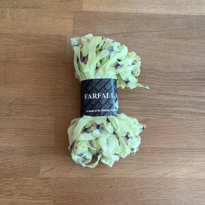 May include: A bundle of light yellow ribbon with a floral pattern, wrapped around a black label that reads "FARFALLA". The ribbon is arranged in a decorative, ruffled style. The background is a wooden surface.