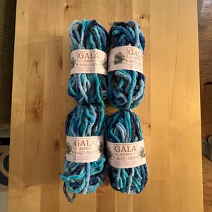 May include: Four skeins of GALA yarns in shades of blue and teal, with a mixed fiber composition. Each skein is wrapped with a label. The yarns are arranged on a light-colored wooden surface.