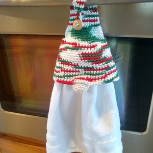 May include: A white kitchen towel with a green, red, and white crocheted hanging loop. The loop has a wooden button and is hanging on a silver oven handle.
