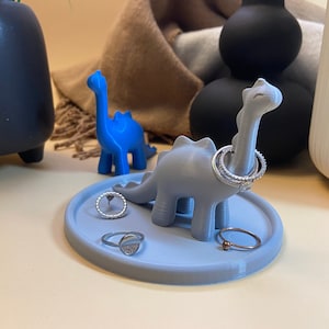 Könnte beinhalten: Ein grauer Dinosaurier-Ringhalter mit einem blauen Dinosaurier im Hintergrund. Der Dinosaurier-Ringhalter hat drei Ringe darauf. Der Ringhalter steht auf einem grauen runden Sockel.