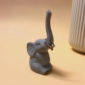Könnte beinhalten: Ein grauer Keramik-Elefanten-Ringständer mit erhobenem Rüssel. Der Elefant hält zwei Ringe in seinem Rüssel.