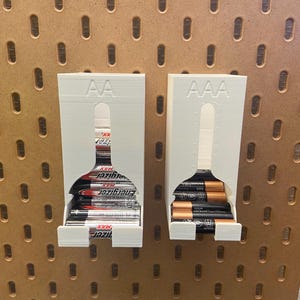 Könnte beinhalten: Zwei weiße Batterie-Organizer aus Kunststoff mit Steckplätzen für AA- und AAA-Batterien. Die Organizer sind an einer Lochwand montiert. Der AA-Organizer ist mit AA-Batterien gefüllt, und der AAA-Organizer ist mit AAA-Batterien gefüllt.