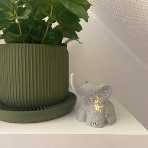 Op de afbeelding: Een groene plantenpot met een geribbeld ontwerp en een bijpassende schotel. Een wit keramiek olifantbeeldje met een lichtje erin staat op een wit rek.