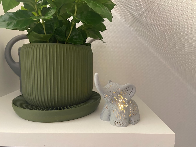 Op de afbeelding: Een groene plantenpot met een geribbeld ontwerp en een bijpassende schotel. Een wit keramiek olifantbeeldje met een lichtje erin staat op een wit rek.