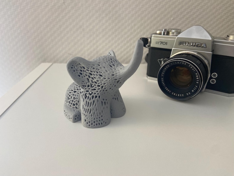 Op de afbeelding: Een grijs, 3D-geprint olifantbeeldje met een gedetailleerd, ingewikkeld ontwerp. De olifant staat op een wit oppervlak met een vintage camera op de achtergrond.