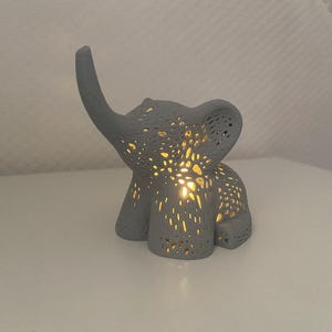 Op de afbeelding: Een grijze keramische olifant nachtlamp met een delicaat, ingewikkeld ontwerp. Het licht is aan en verlicht het lichaam van de olifant.