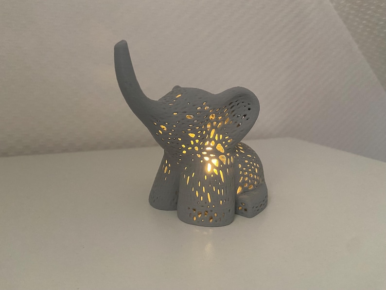 Op de afbeelding: Een grijze keramische olifant nachtlamp met een delicaat, ingewikkeld ontwerp. Het licht is aan en verlicht het lichaam van de olifant.