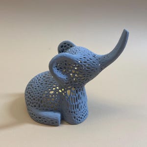 Op de afbeelding: Een lichtblauw, 3D-geprint olifantenbeeldje met een gedetailleerd, ingewikkeld, traliewerk-achtig ontwerp. De olifant heeft zijn slurf omhoog in de lucht.