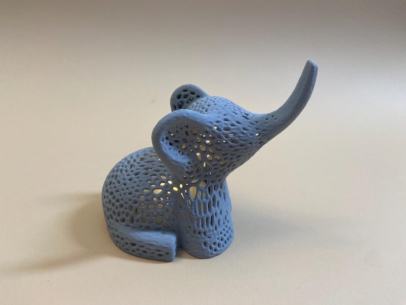 Op de afbeelding: Een lichtblauw, 3D-geprint olifantenbeeldje met een gedetailleerd, ingewikkeld, traliewerk-achtig ontwerp. De olifant heeft zijn slurf omhoog in de lucht.