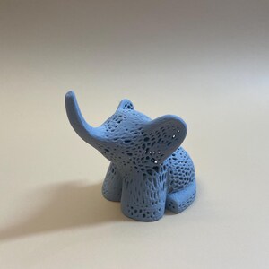 Op de afbeelding: Een lichtblauw, 3D-geprint olifantbeeldje met een delicaat, kant-achtig ontwerp. De olifant zit met zijn slurf omhoog.