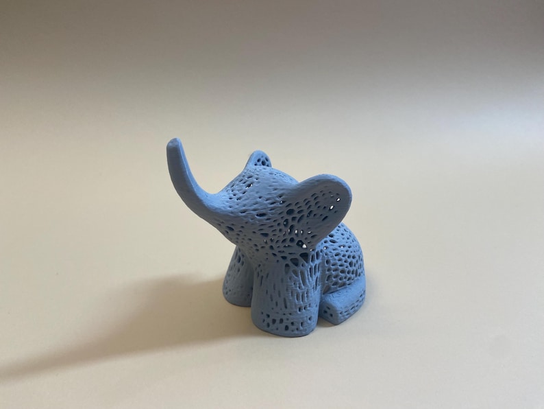 Op de afbeelding: Een lichtblauw, 3D-geprint olifantbeeldje met een delicaat, kant-achtig ontwerp. De olifant zit met zijn slurf omhoog.