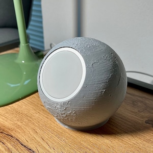 Könnte beinhalten: Eine graue mondförmige Lampe mit einem weißen Licht im Inneren. Die Lampe steht auf einer Holzoberfläche.