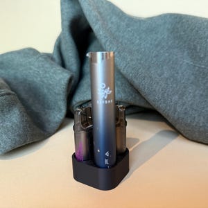 Könnte beinhalten: Ein schwarzer Ladeständer mit drei Vape-Pods. Der mittlere Pod ist silberfarben mit dem aufgedruckten Wort "ELUXBAR". Die beiden anderen Pods sind schwarz und lila.