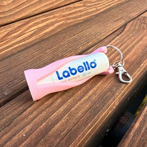 Könnte beinhalten: Eine weiße Labello Lippenbalsam-Tube mit blauem Text und einer rosa Halterung, die an einem silbernen Schlüsselanhänger befestigt ist. Der Schlüsselanhänger hat einen Karabinerverschluss. Die Artikel befinden sich auf einer Holzoberfläche.