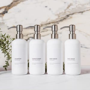May include: Four white, refillable pump bottles labeled "Shampoo", "Conditioner", "Body Wash", and "Face Wash" with silver-colored pumps. A small potted plant is to the left. The background is a marble-patterned wall.