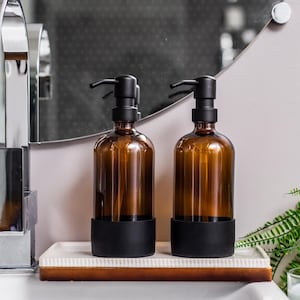 May include: Two amber glass soap dispensers with black pumps and black rubber bases on a white rectangular tray. The dispensers are in a bathroom setting with a chrome faucet and a green plant.