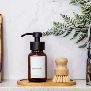 May include: A brown glass bottle with a black pump dispenser filled with hand soap. The bottle has a white label with the text "HAND SOAP" and "GARDEN & HANDS". The bottle is sitting on a wooden tray with a wooden brush next to it.