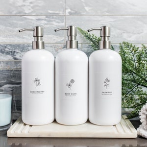 May include: Three white pump bottles with black labels and simple floral designs. The bottles are labelled "Conditioner", "Body Wash", and "Shampoo". The bottles are sitting on a white marble tray.