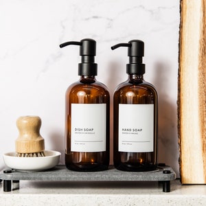 May include: Two amber glass bottles with black pump tops and white labels. The bottles are labelled "Dish Soap" and "Hand Soap". The bottles are on a grey tray with a wooden brush and a black cloth.