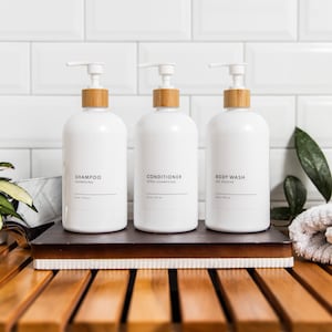 May include: Three white pump bottles with bamboo lids and labels reading "Shampoo", "Conditioner", and "Body Wash" on a wooden shelf.
