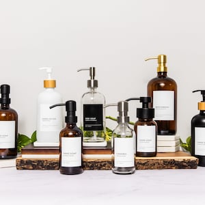 May include: A collection of eight amber glass bottles with black and gold pump tops. The bottles are labelled with white text on black labels. The labels read "Hand Soap", "Hand Lotion", "Dish Soap", "Mouthwash", "Caramel", and "Hand Soap". The bottles are arranged on a wooden surface with green plants in the background.