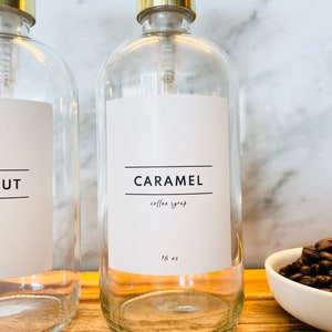 May include: Clear glass syrup bottles with gold-colored pump tops. One bottle has a label that reads "CARAMEL coffee syrup 16 oz." and the other reads "HAZELNUT coffee syrup 16 oz." A small white bowl of coffee beans is in the background.
