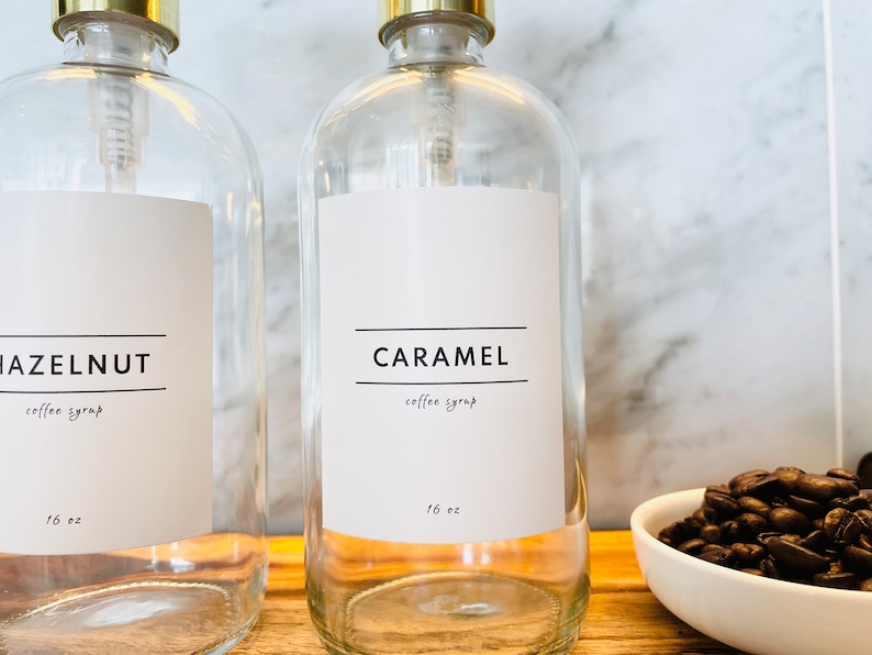 May include: Clear glass syrup bottles with gold-colored pump tops. One bottle has a label that reads "CARAMEL coffee syrup 16 oz." and the other reads "HAZELNUT coffee syrup 16 oz." A small white bowl of coffee beans is in the background.