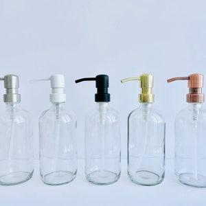 May include: Five clear glass soap dispenser bottles with different pump finishes: silver, white, black, gold, and copper. The bottles are oval-shaped and have a simple, modern design, suitable for bathroom or kitchen use.