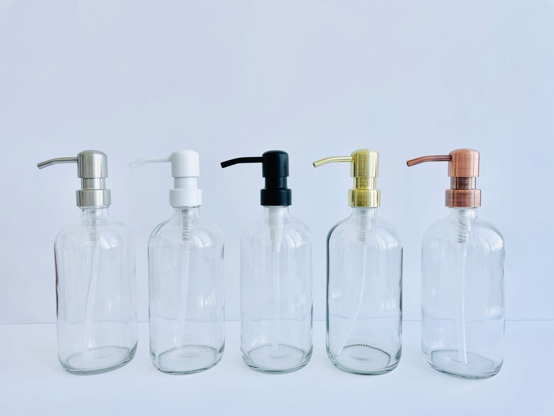 May include: Five clear glass soap dispenser bottles with different pump finishes: silver, white, black, gold, and copper. The bottles are oval-shaped and have a simple, modern design, suitable for bathroom or kitchen use.