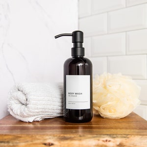 May include: A black pump bottle with a white label that reads "BODY WASH - OIL SOYBEAN" on a wooden surface with a white towel and a yellow bath sponge.