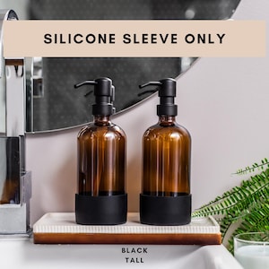 Peut inclure: Deux flacons en verre ambré avec des pompes noires et des manchons en silicone noir. Les flacons sont étiquetés "BLACK TALL" et sont posés sur un plateau blanc. Le texte "SILICONE SLEEVE ONLY" est affiché au-dessus des flacons.