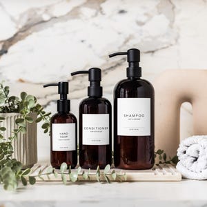 May include: Three dark brown pump bottles labelled "Hand Soap", "Conditioner", and "Shampoo" with white labels. A rolled white towel and a beige decorative object are next to the bottles. A potted plant with green leaves is on the left.