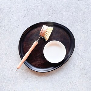 May include: A wooden dish brush with a wooden handle and a natural bristle brush head is on a dark wooden tray with a small white bowl.