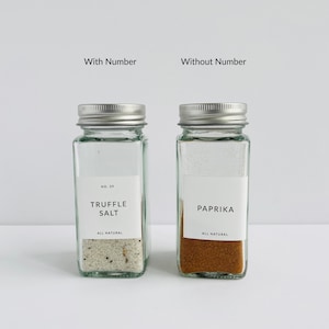 以下が含まれることがあります： 銀色の蓋が付いた2つの透明なガラス製スパイスジャー。1つは「Truffle Salt」、もう1つは「Paprika」とラベル付けされています。どちらのジャーにも、黒い文字の白いラベルが付いています。「Truffle Salt」ジャーには「No. 39」のラベルが付いています。