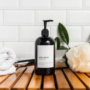 May include: Black pump bottle with a white label that reads "BODY WASH GEL DOUCHE" on a wooden surface with a white washcloth and a white bath pouf.