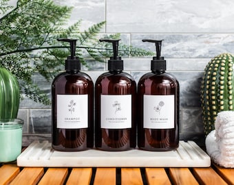 May include: Three brown glass bottles with pumps, labeled "Shampoo", "Conditioner", and "Body Wash". The bottles are on a white marble tray, with a green cactus and a white towel in the background.
