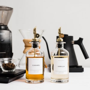 May include: Two glass bottles with gold spouts and labels that say "Caramel" and "Vanilla". The bottles are filled with a light brown and a light yellow liquid. The bottles are on a white surface.