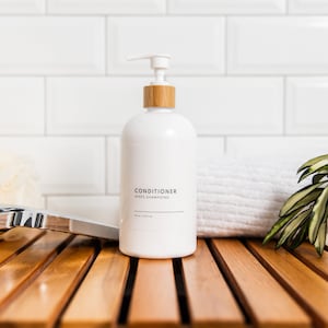 May include: White pump bottle with a wooden top and a label that reads "CONDITIONER APRES SHAMPOOING 10 fl oz (295 ml)". The bottle is sitting on a wooden surface with a white towel and a green plant in a white pot.